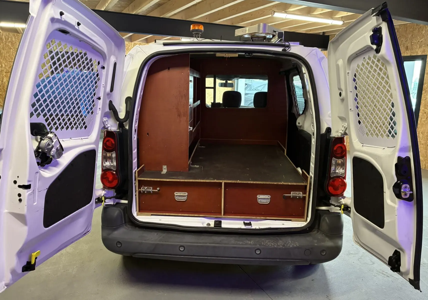 Vue arrière ouverte d’un Citroën Berlingo blanc 2016 avec aménagement bois intérieur et portes battantes ajourées.