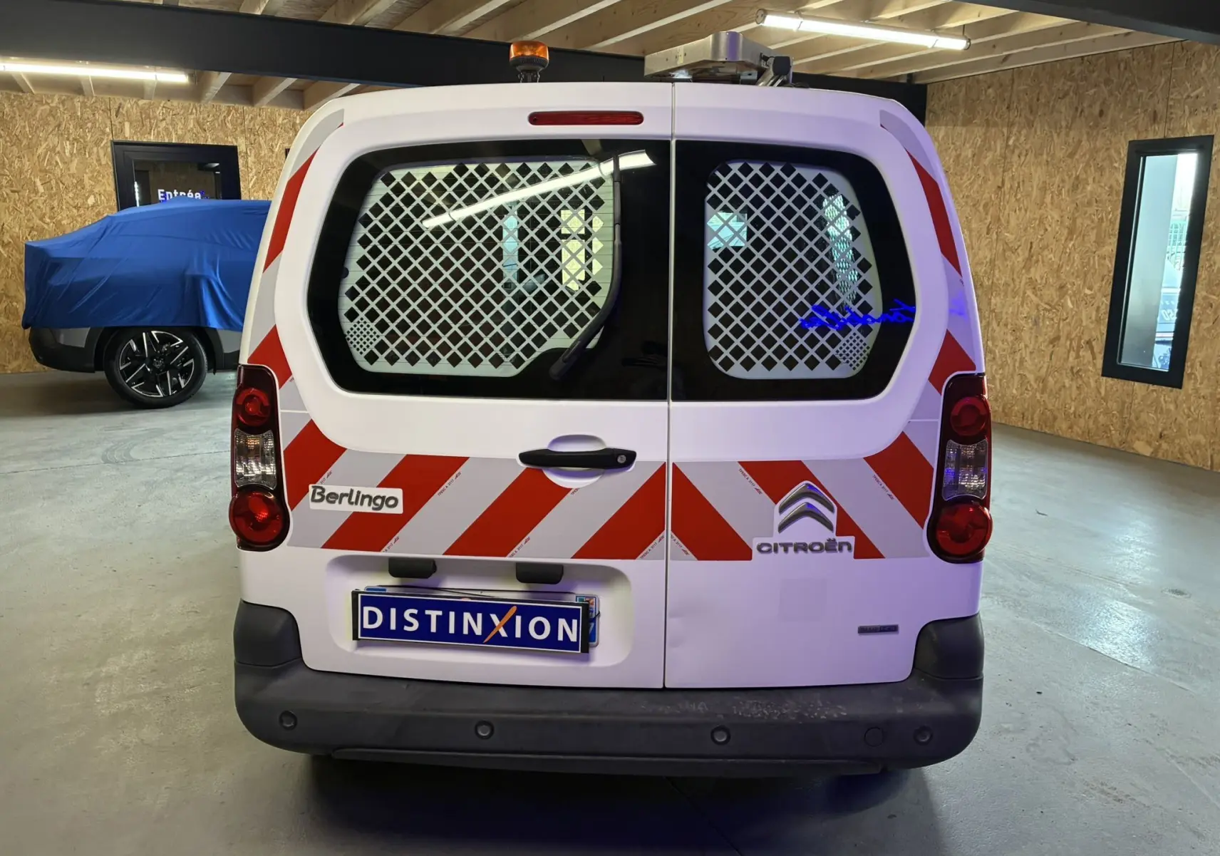 Vue arrière d'un Citroën Berlingo blanc 2016 avec bandes rouges diagonales et grille de protection sur les vitres arrière.