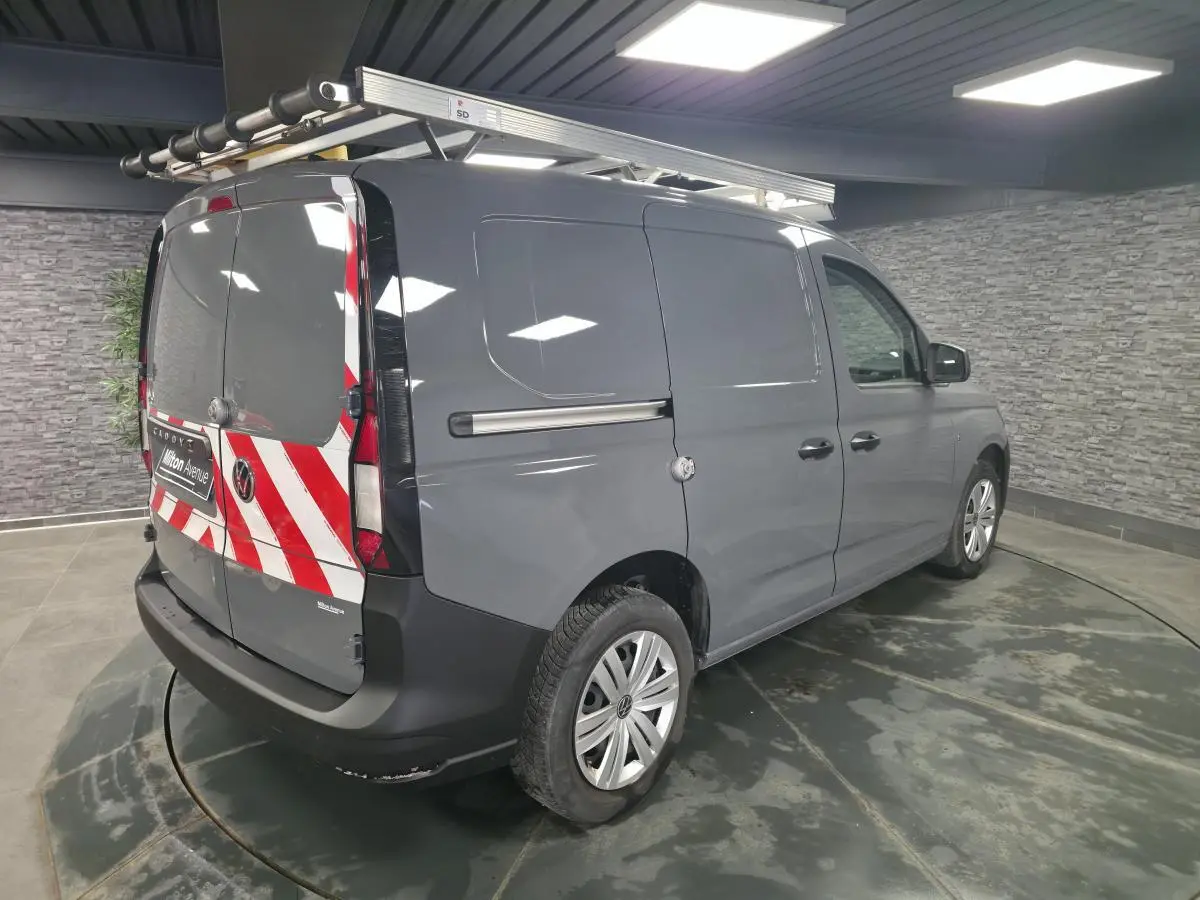 Vue 3/4 arrière droite du Volkswagen Caddy Cargo gris avec galerie de toit et marquage rouge et blanc sur portes arrière.