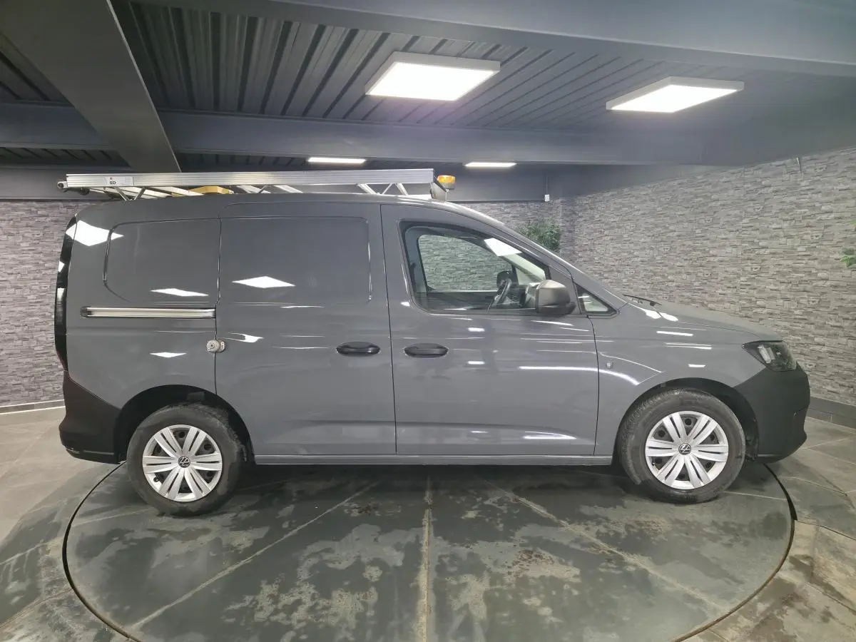 Vue de profil droit du Volkswagen Caddy Cargo gris avec galerie de toit et jantes acier, dans un showroom intérieur.