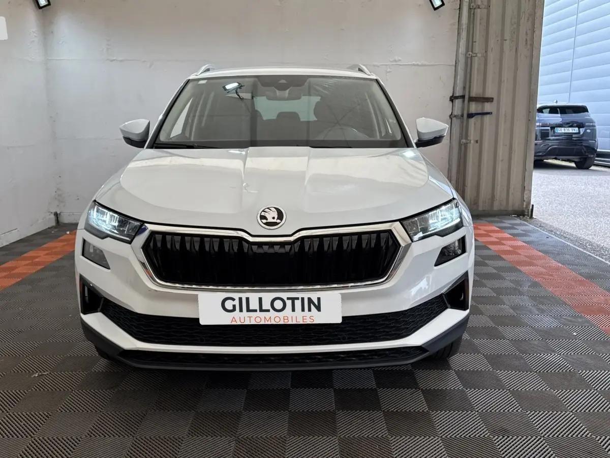 Vue de face du SUV blanc SKODA KAROQ 2024 avec calandre noire et phares LED allumés dans un garage.