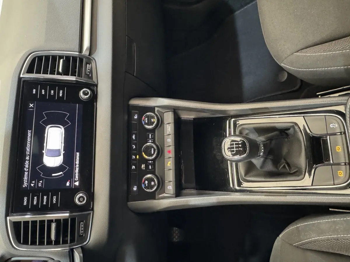 Vue intérieure centrée sur la console centrale du Skoda Karoq blanc, avec levier de vitesses manuel et écran tactile affichant l'aide au stationnement.