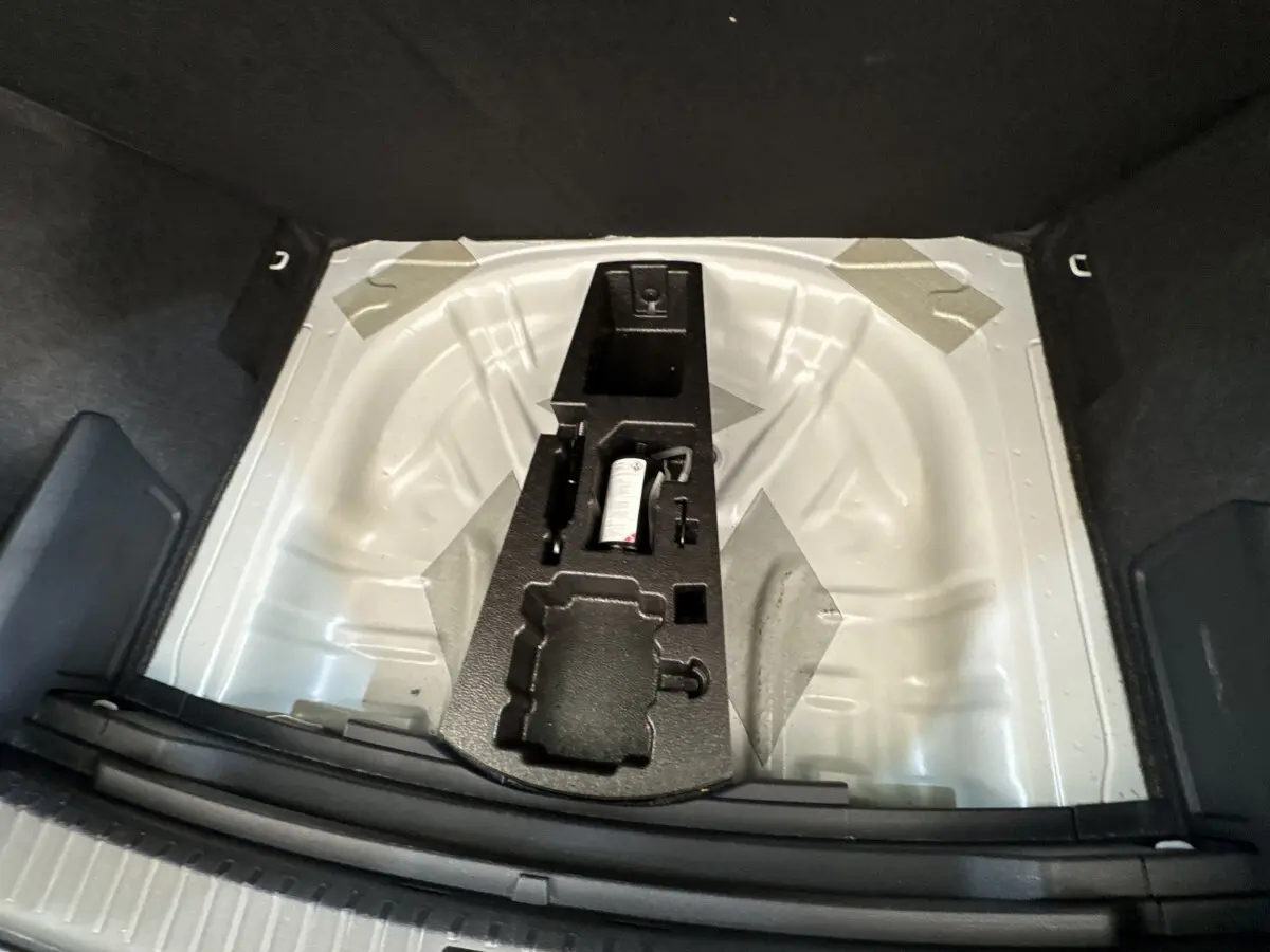 Vue plongeante sur le coffre ouvert d’un Skoda Karoq blanc 2024, montrant le rangement pour roue de secours et outils.