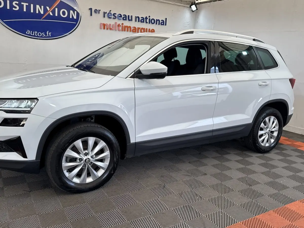 Vue de profil côté gauche d'un Skoda Karoq blanc 2024 avec jantes alliage et barres de toit noires en showroom.