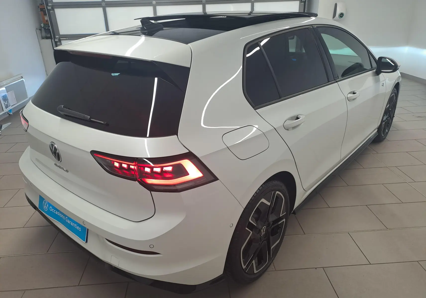 Vue 3/4 arrière droite d'une Volkswagen Golf blanche nacrée avec toit noir et feux arrière LED allumés dans un garage.
