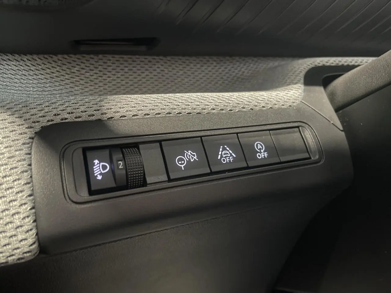 Gros plan sur les commandes d’assistance et réglage des phares à gauche du tableau de bord d’une Citroën C3 gris foncé.