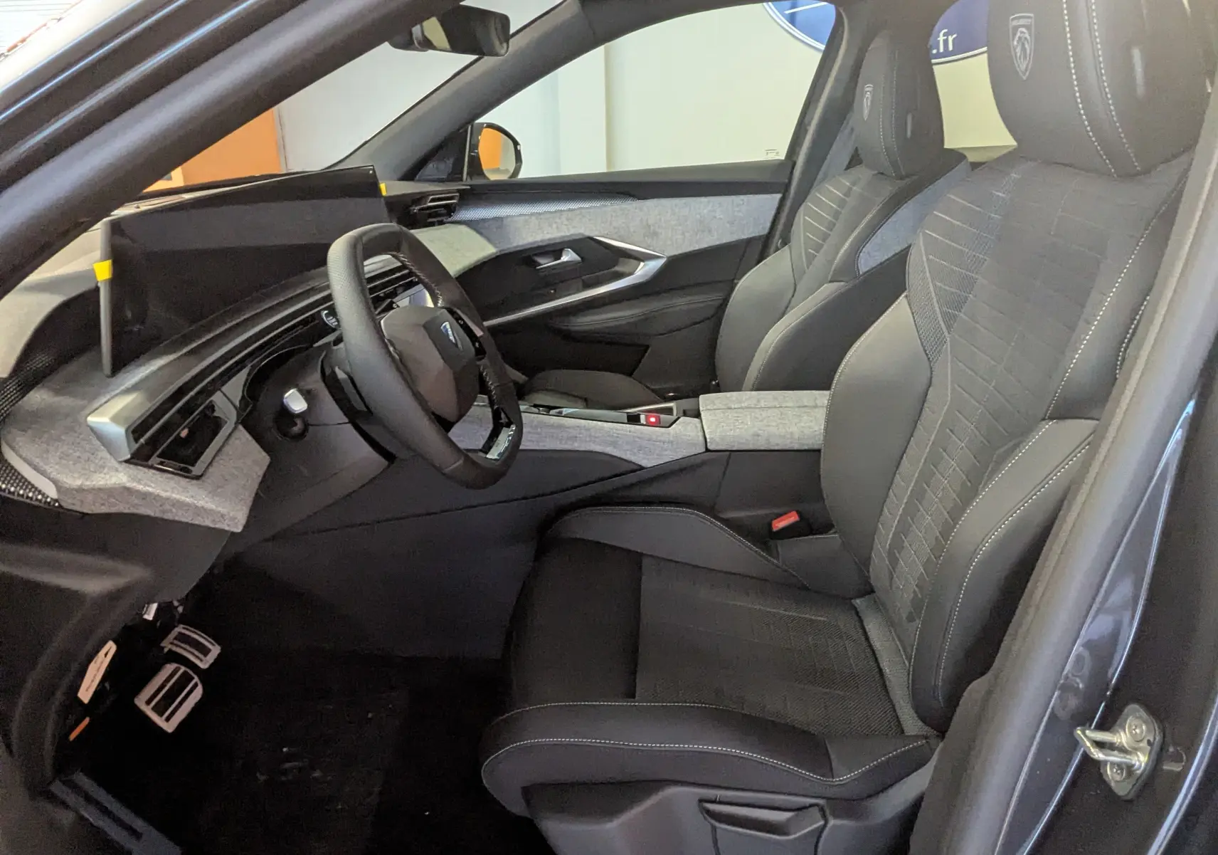 Vue intérieure côté conducteur du Peugeot 3008 Hybrid 145 GT 2025, sièges tissu/tepp gris avec surpiqûres et pédalier alu visible.