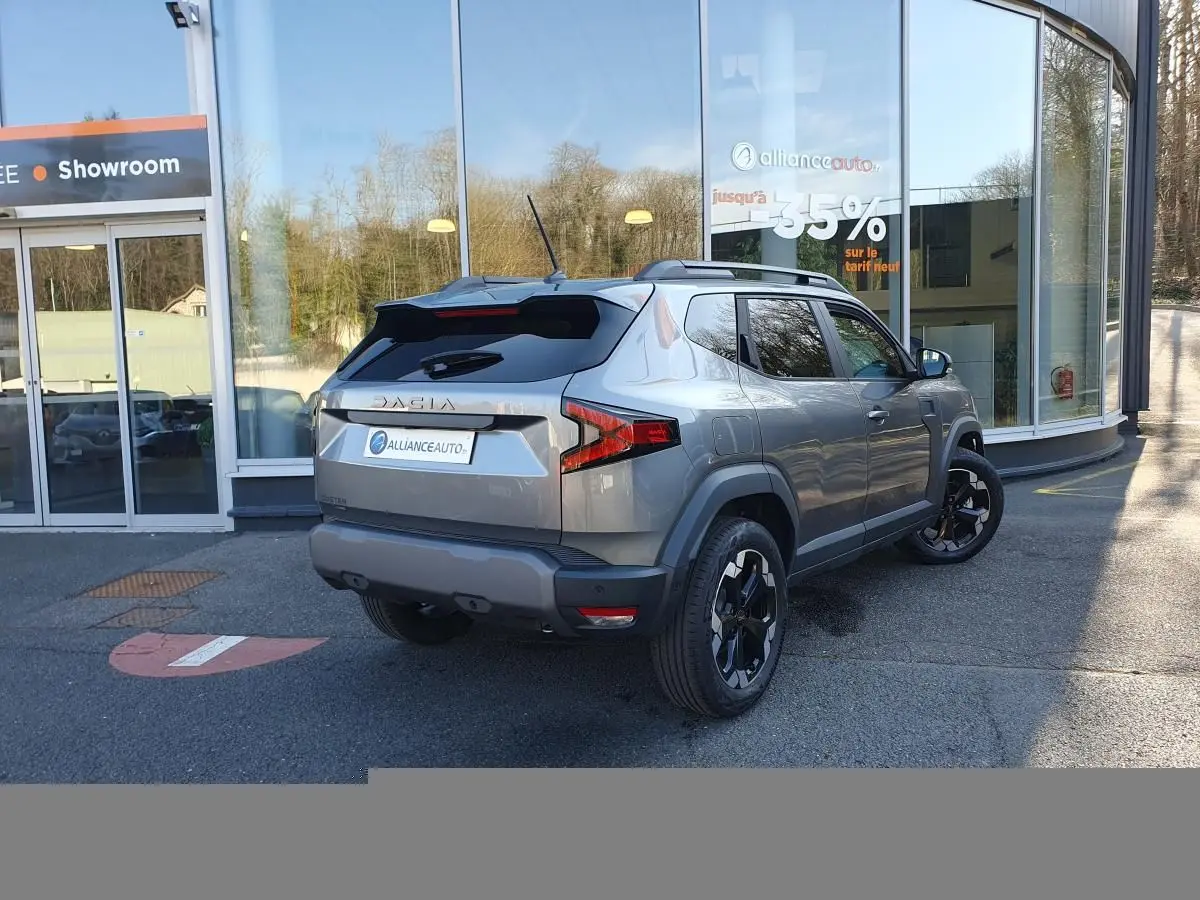 Vue 3/4 arrière droite du Dacia Duster 2026 gris schiste avec jantes alliage noires et toit noir, devant un showroom.