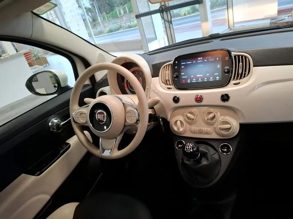 Vue intérieure côté conducteur de la FIAT 500 blanche avec tableau de bord beige et levier de vitesses manuel visible.