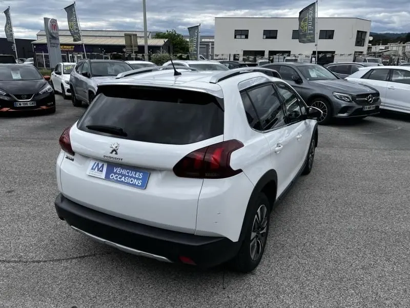 Peugeot 2008 blanc vue 3/4 arrière droit, avec vitres teintées et barres de toit, sur parking de voitures d'occasion.