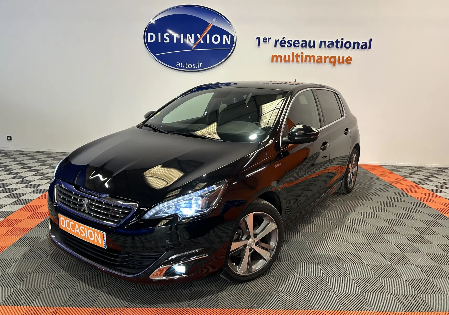 Peugeot 308 noire vue 3/4 avant droit avec phares allumés et jantes alliage dans showroom.