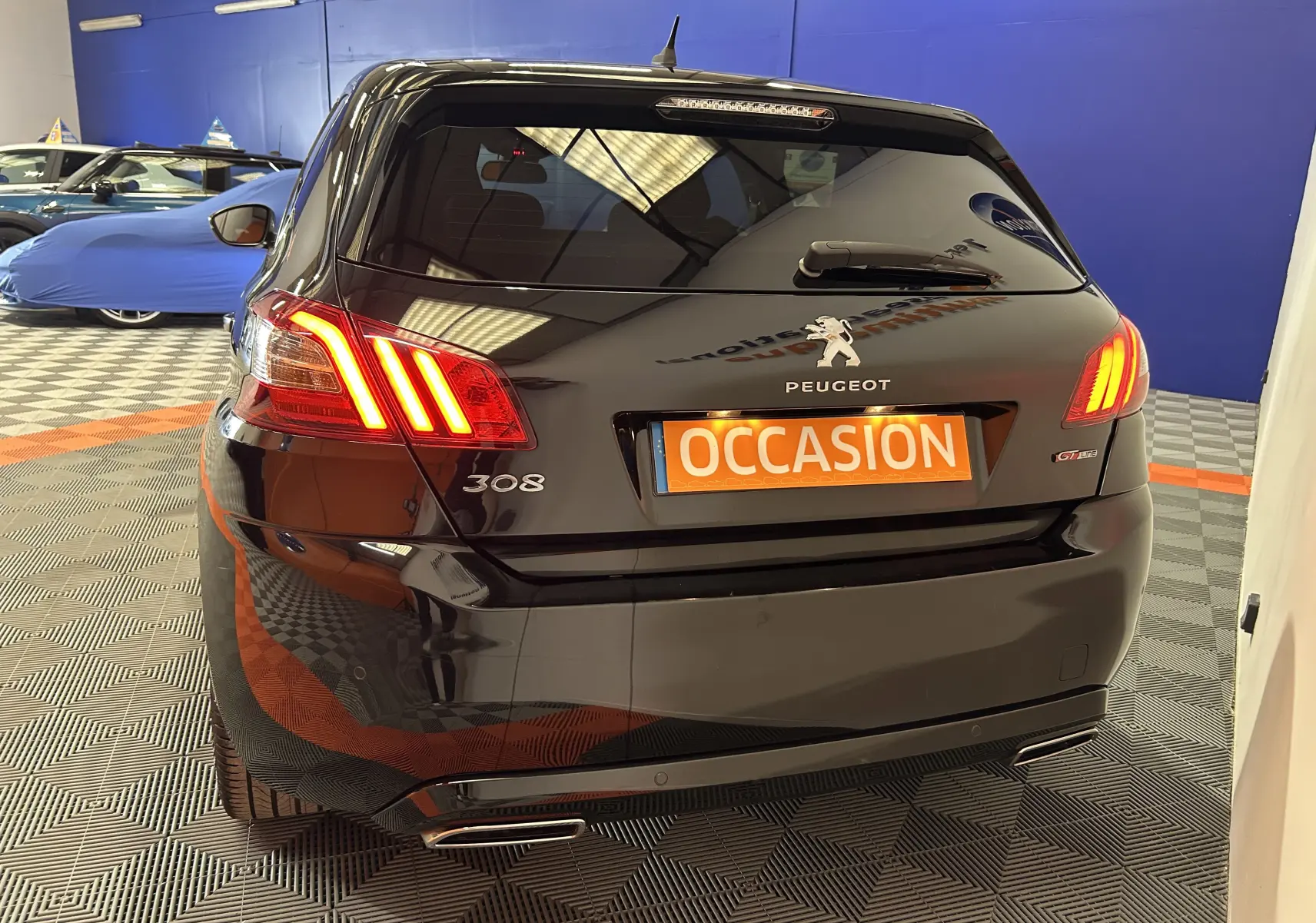Vue 3/4 arrière d'une Peugeot 308 noire avec feux allumés et plaque orange "OCCASION" en intérieur showroom.