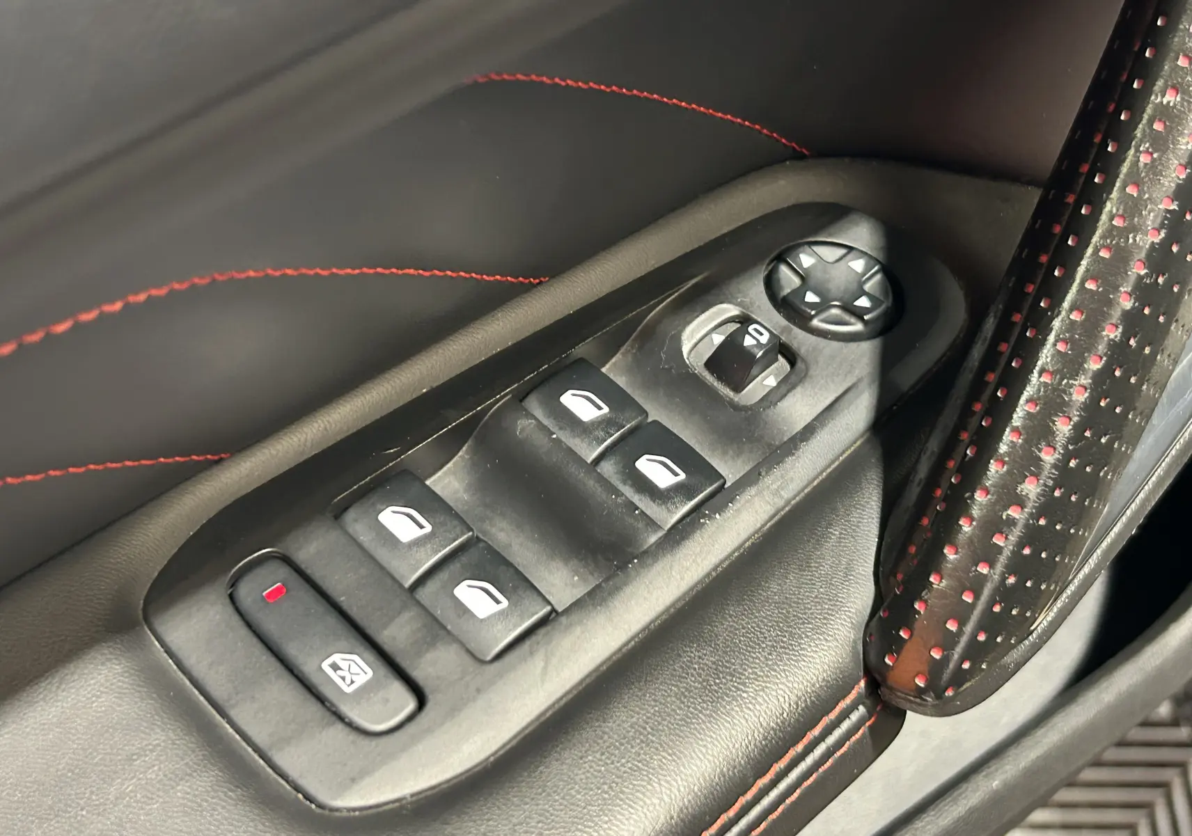 Gros plan sur les commandes de lève-vitres et réglage des rétroviseurs côté conducteur, avec surpiqûres rouges sur cuir noir.