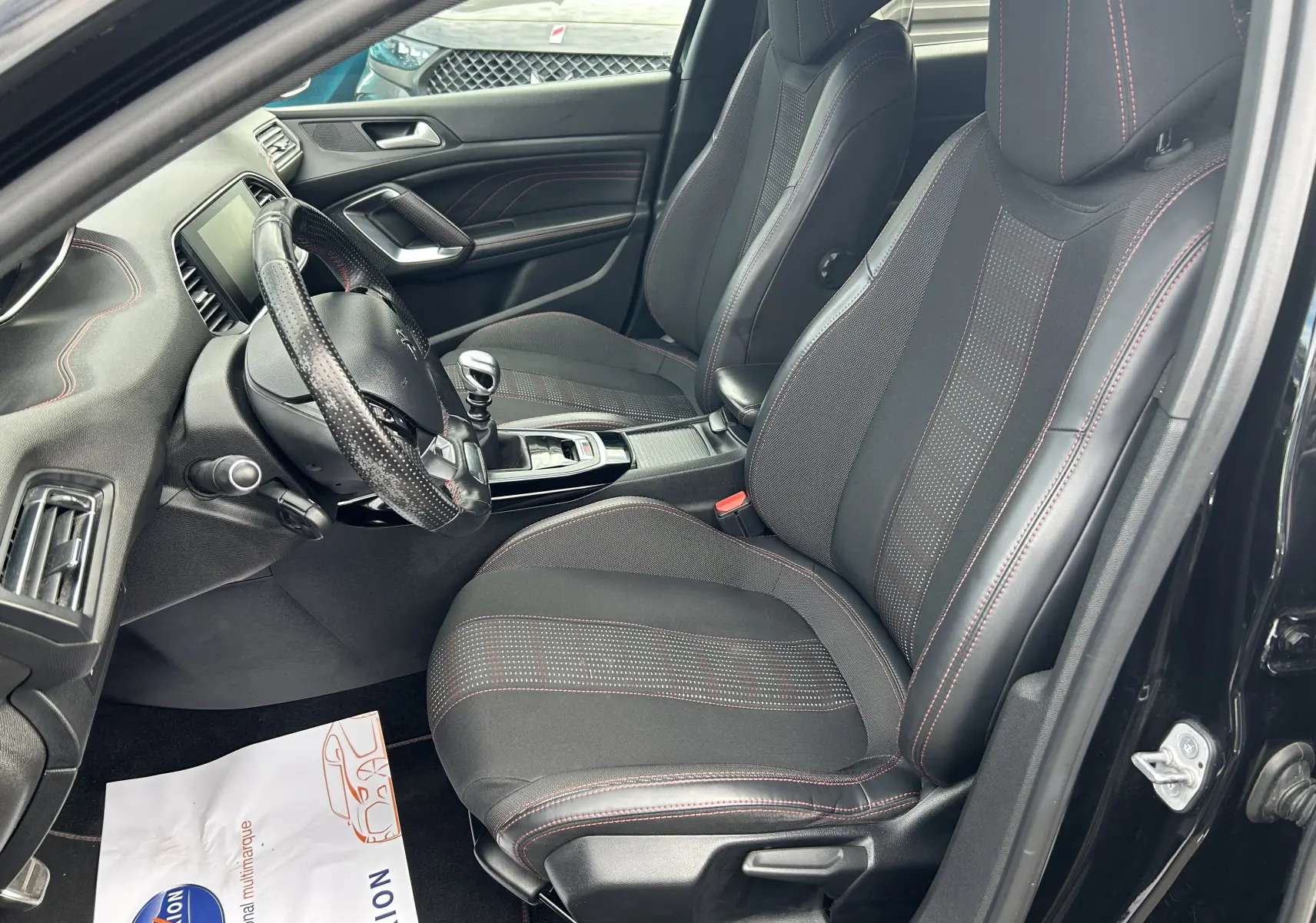 Vue intérieure côté conducteur de la Peugeot 308 noire, avec sièges sport noirs et coutures rouges visibles.