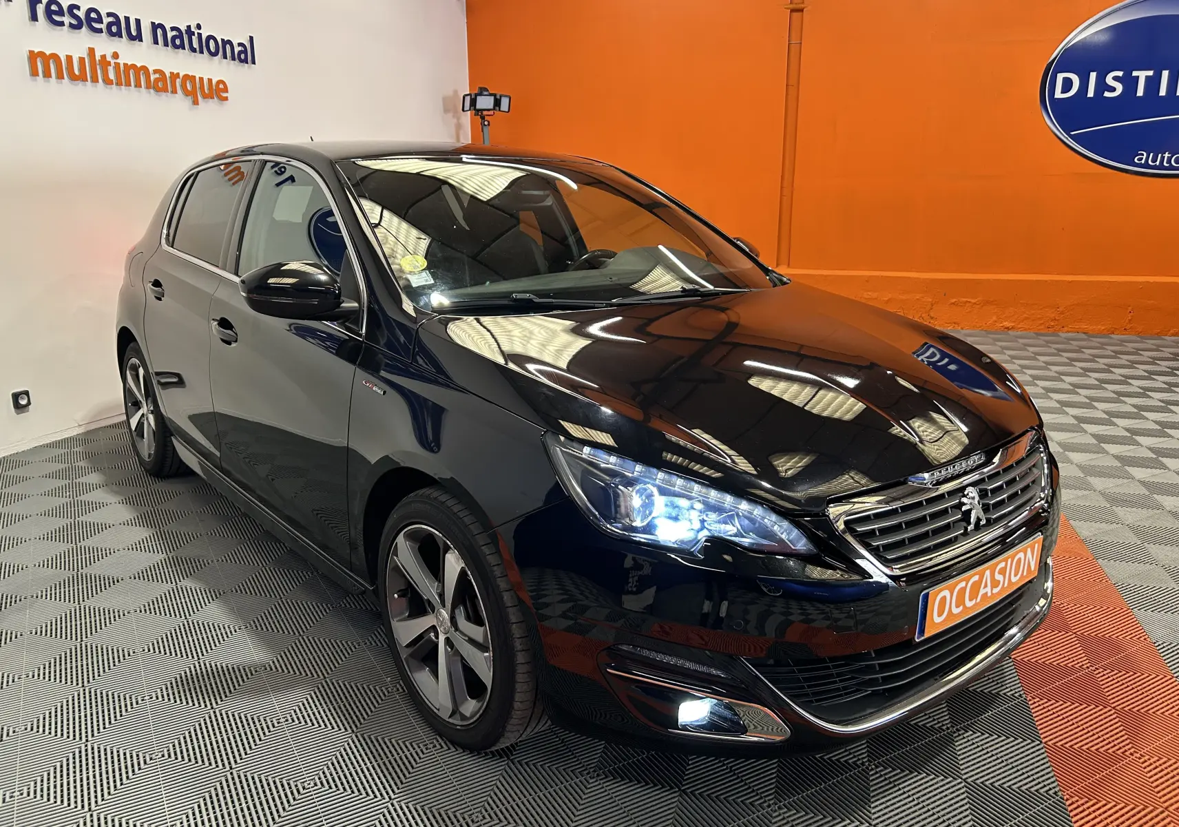 Peugeot 308 noire en 3/4 avant droit avec phares allumés et jantes alliage bi-ton dans un showroom.