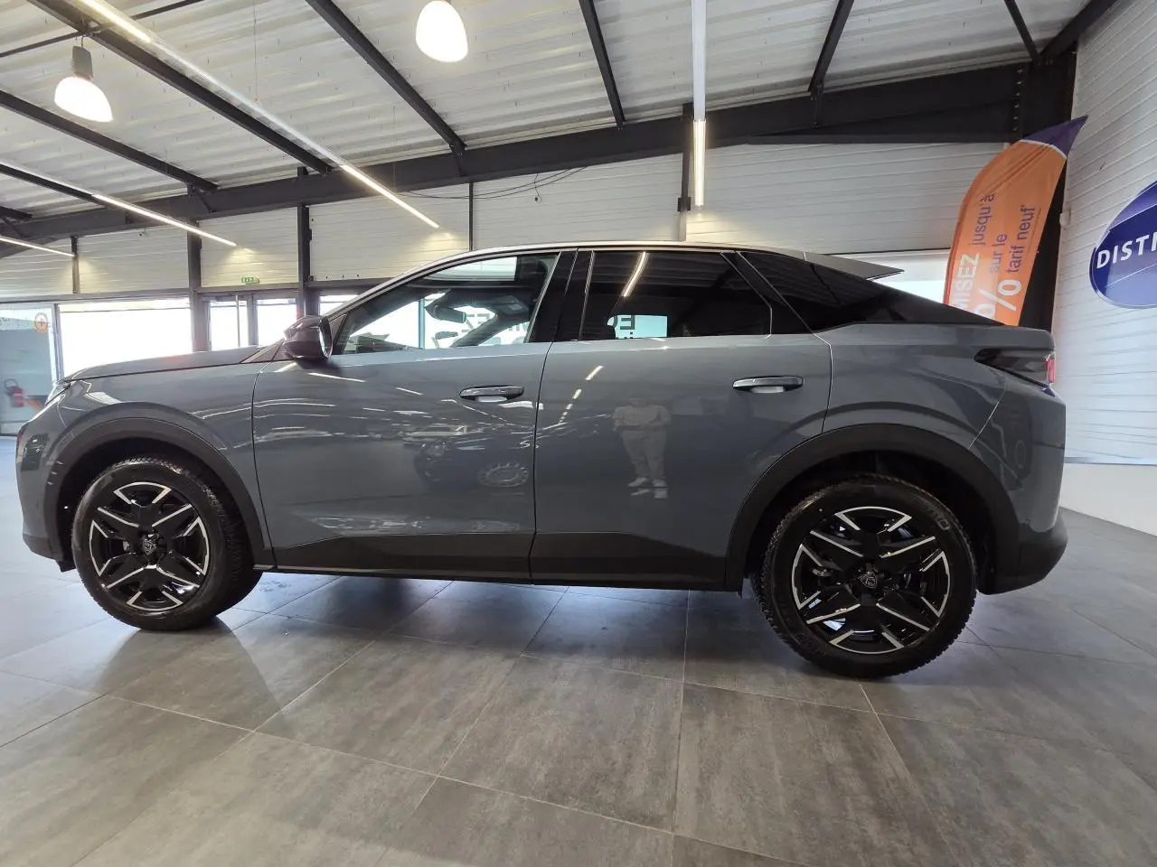 Vue latérale droite d’un Peugeot 3008 hybride 2025 gris métallisé avec jantes alliage noires et toit noir brillant en intérieur showroom.