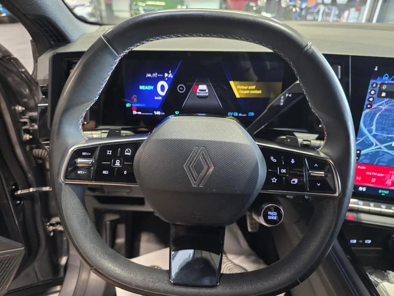 Vue rapprochée du volant en textile noir avec insert Alcantara et logo Renault, tableau de bord numérique en arrière-plan.
