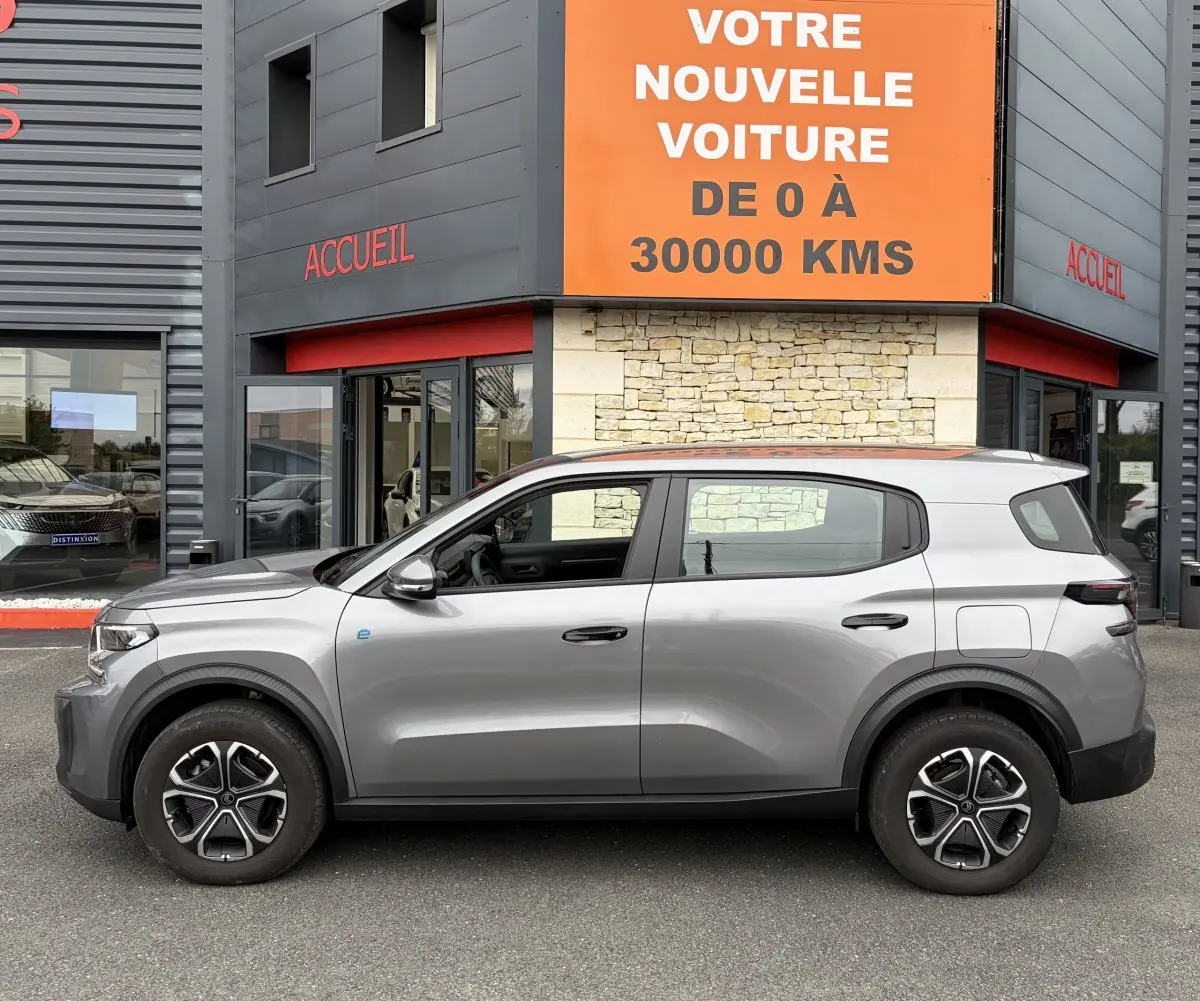 Vue de profil côté gauche d'une Citroën C3 Aircross électrique gris foncé garée devant un bâtiment avec panneau orange.