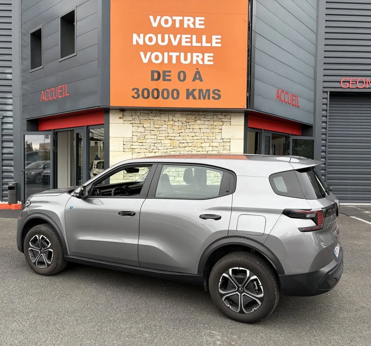 Vue de profil côté gauche d'un Citroën C3 Aircross électrique gris foncé garé devant un bâtiment avec panneau orange.