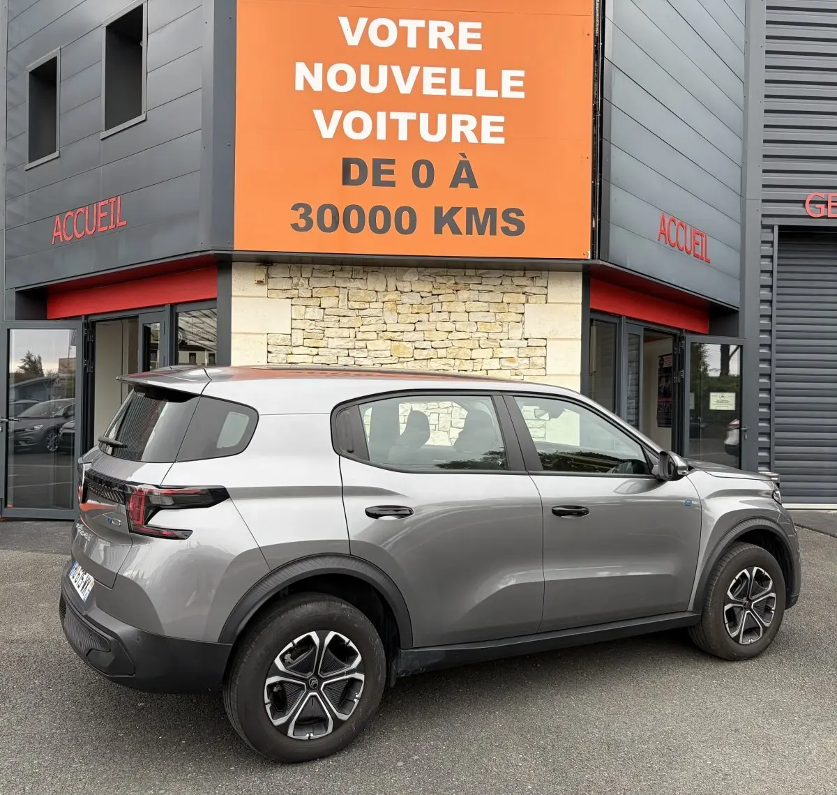 Vue latérale droite d'un Citroën C3 Aircross électrique gris foncé garé devant un bâtiment avec enseigne orange.