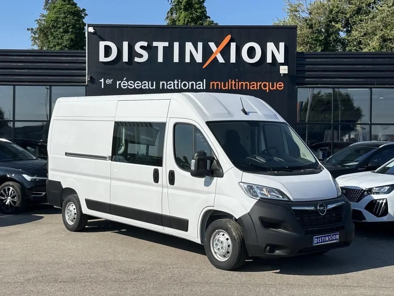 Fourgon blanc Opel Movano L3H2 en 3/4 avant droit, avec porte latérale vitrée et crochet d'attelage visible.