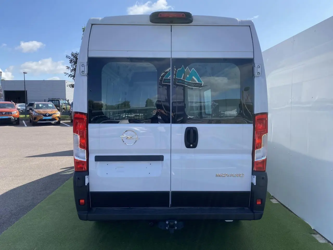 Vue arrière d'un Opel Movano Fourgon blanc avec portes vitrées battantes et crochet d'attelage visible.