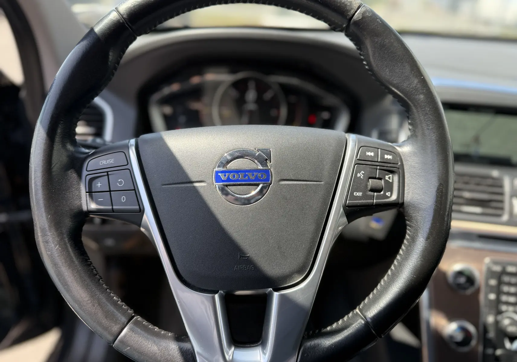 Vue rapprochée du volant en cuir noir du Volvo XC60 gris foncé, avec commandes audio et tableau de bord en arrière-plan.