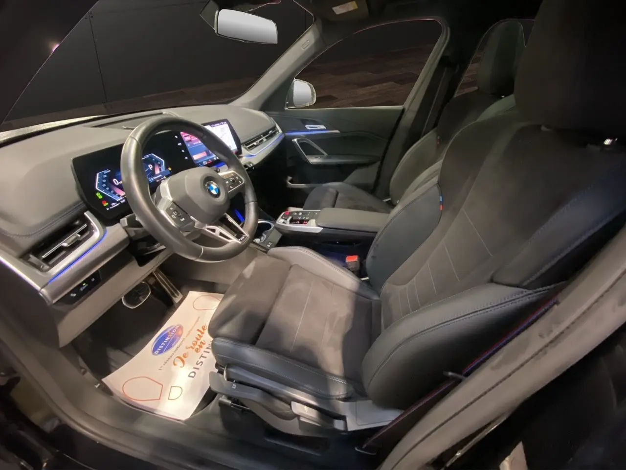 Intérieur BMW X1 2025 vu côté conducteur, sièges noirs M Sport avec tableau de bord digital et éclairage d'ambiance bleu.