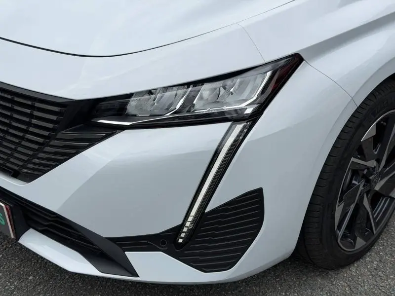 Gros plan sur l'avant droit blanc Okénite d'une Peugeot 308 2025, mettant en valeur le phare LED et la jante alu noire.