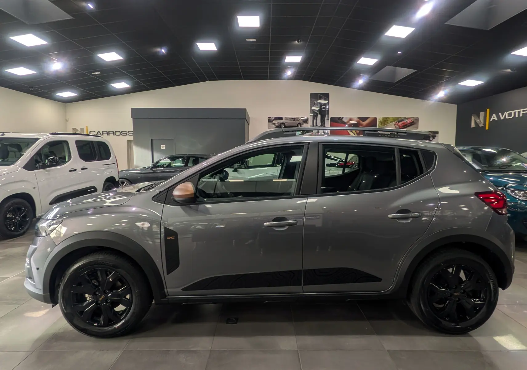 Profil côté gauche d'une Dacia Sandero Stepway TCe 110 Gris Schiste avec jantes noires et barres de toit en showroom.