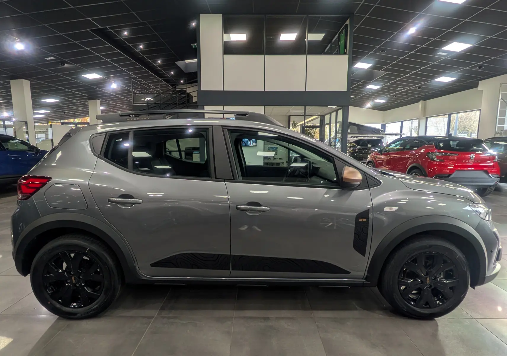 Profil droit d'une Dacia Sandero Stepway TCe 110 Gris Schiste avec jantes noires et détails noirs sur les portes, en showroom.