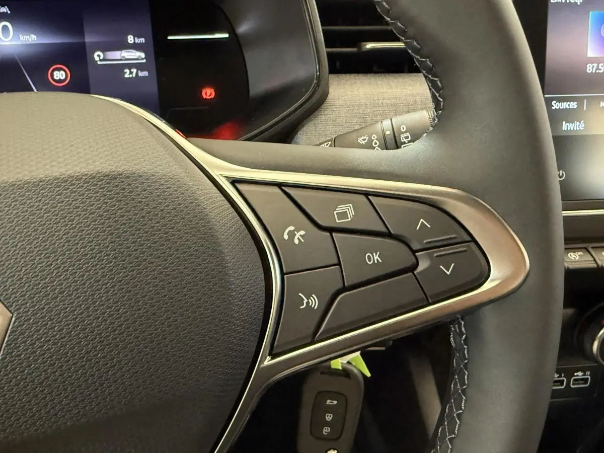 Gros plan sur le volant cuir noir de la Renault Clio 2025, avec commandes multifonctions et tableau de bord digital en arrière-plan.