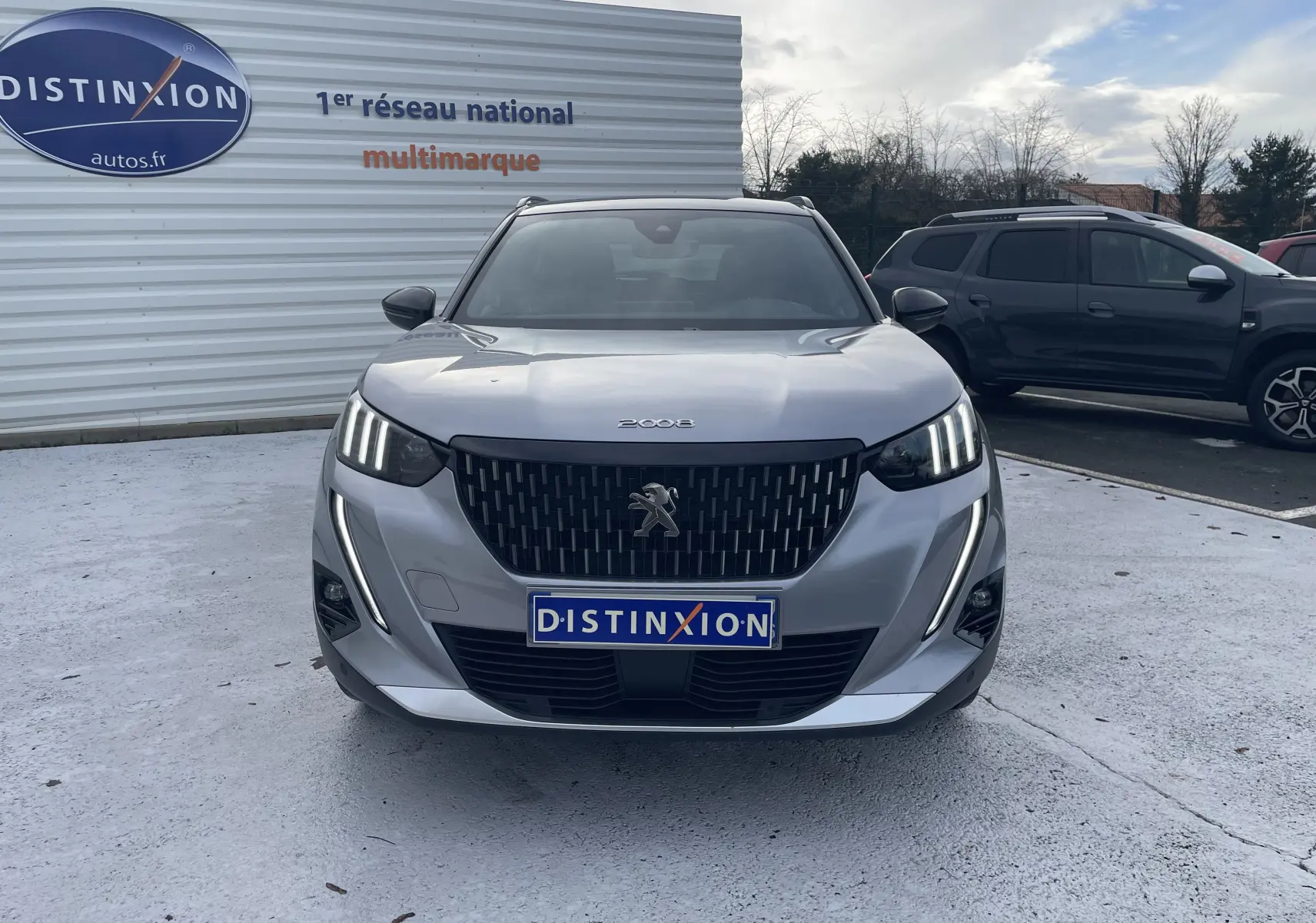 Vue frontale d'un Peugeot 2008 gris Artense avec calandre noire et feux LED distinctifs allumés, sur parking extérieur.