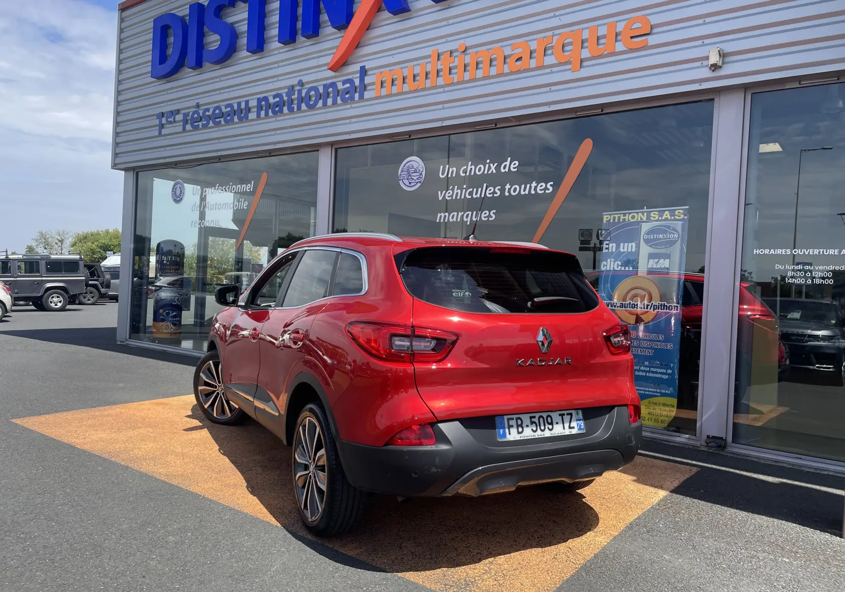 Vue 3/4 arrière droite d’un Renault Kadjar rouge flamme garé devant un showroom multimarque avec jantes alliage bicolores.