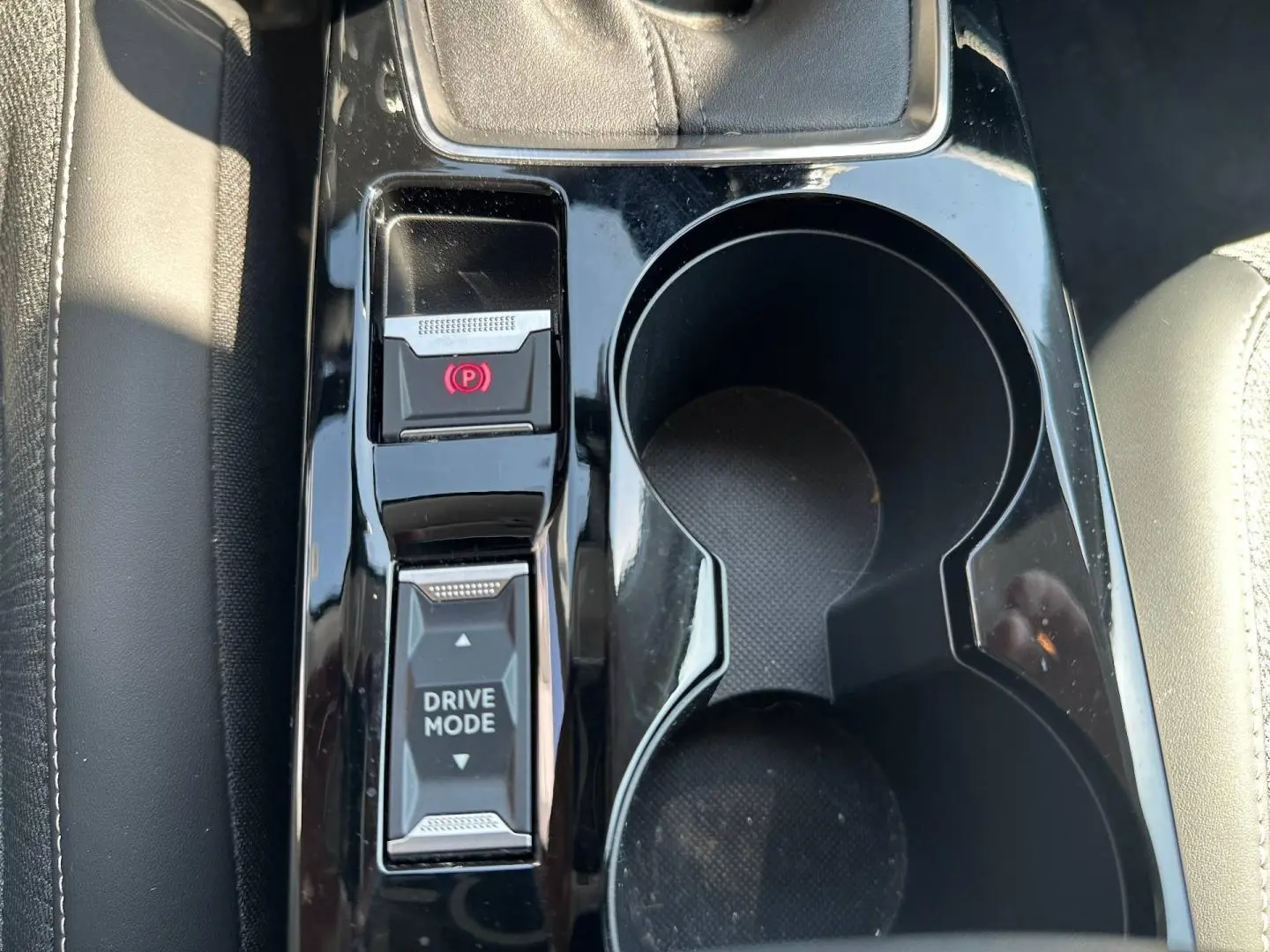 Gros plan sur la console centrale noire brillante avec bouton frein à main électrique et sélecteur Drive Mode, sièges gris visibles.