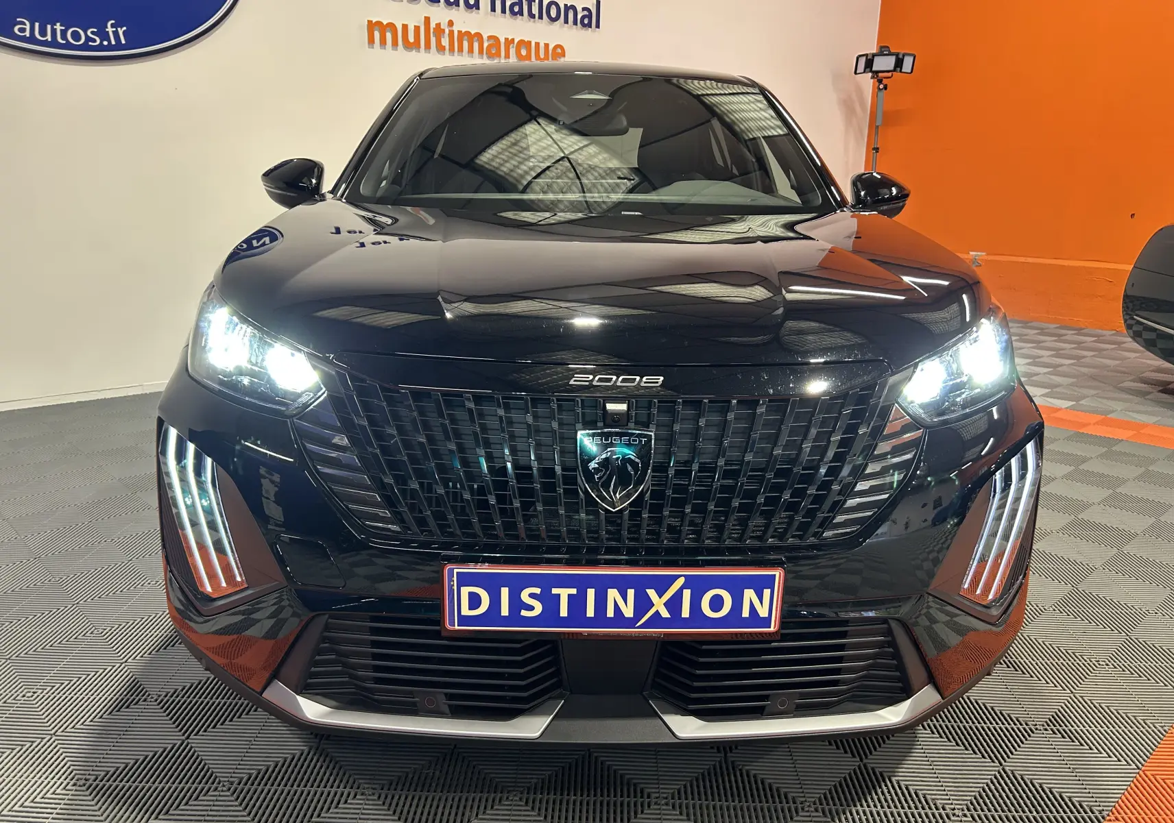 Vue frontale d'un Peugeot 2008 noir avec phares allumés et calandre imposante, en intérieur showroom.