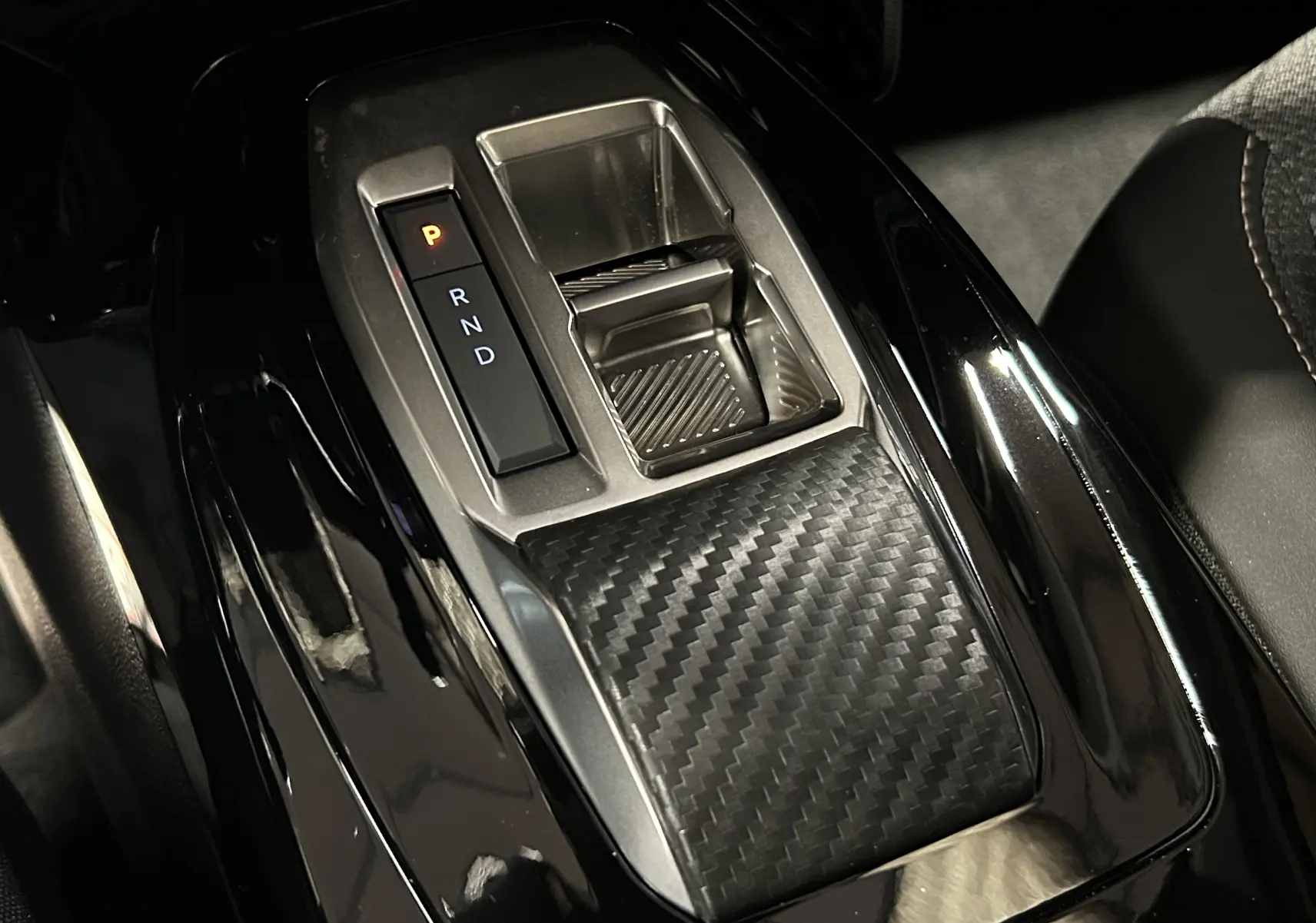 Gros plan sur la console centrale noire brillante du Peugeot 2008 II Hybrid avec levier de vitesses automatique et finition carbone.