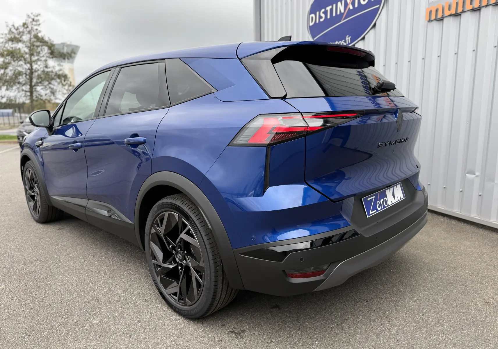 Renault Symbioz bleu Iron vu en 3/4 arrière droit, avec jantes noires et hayon électrique visible.