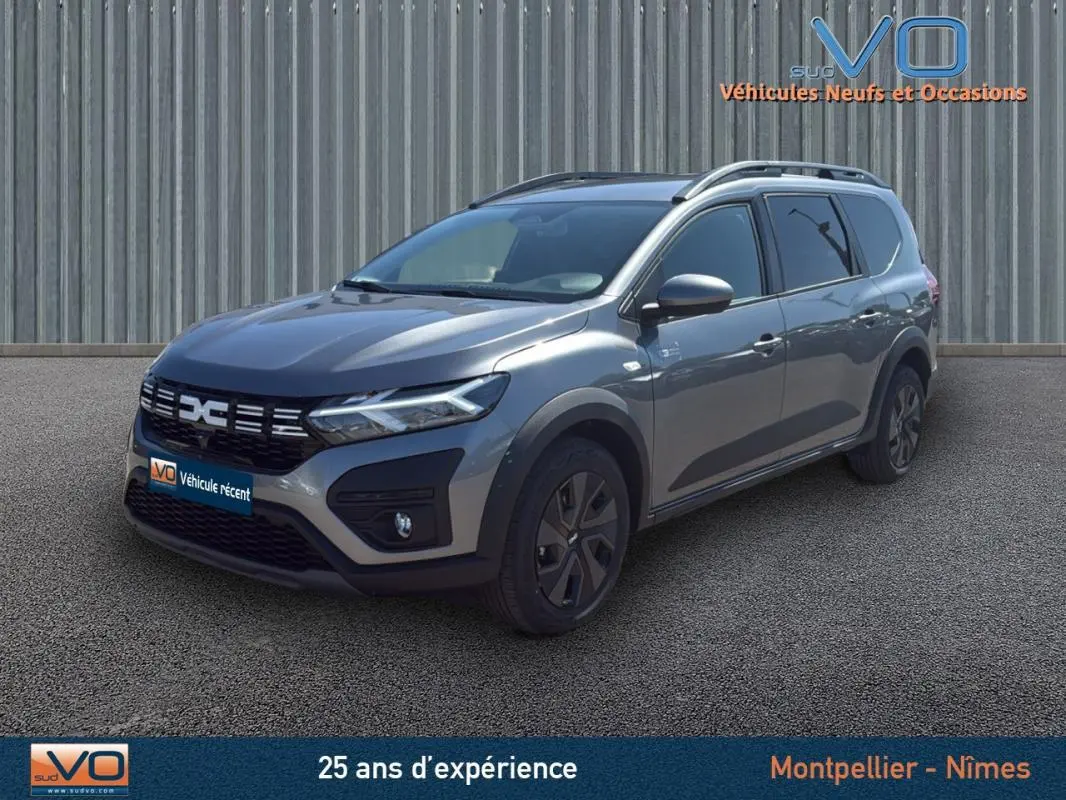 Vue 3/4 avant droite d'un Dacia Jogger gris 2025 avec barres de toit et jantes noires, stationné devant un mur métallique.