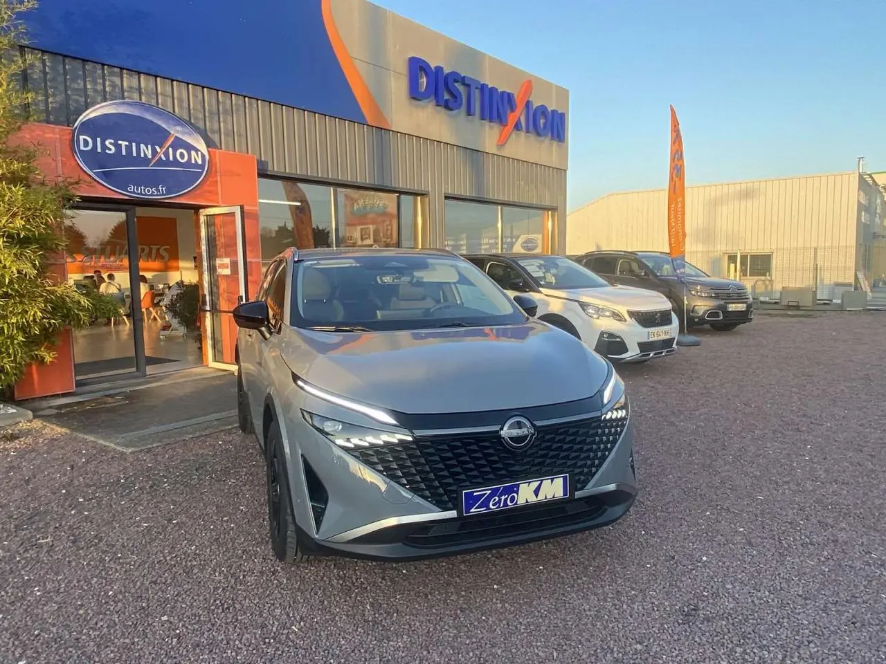 Vue 3/4 avant d'un Nissan Qashqai gris ceramic 2025 garé devant un concessionnaire avec plaque "Zéro KM".