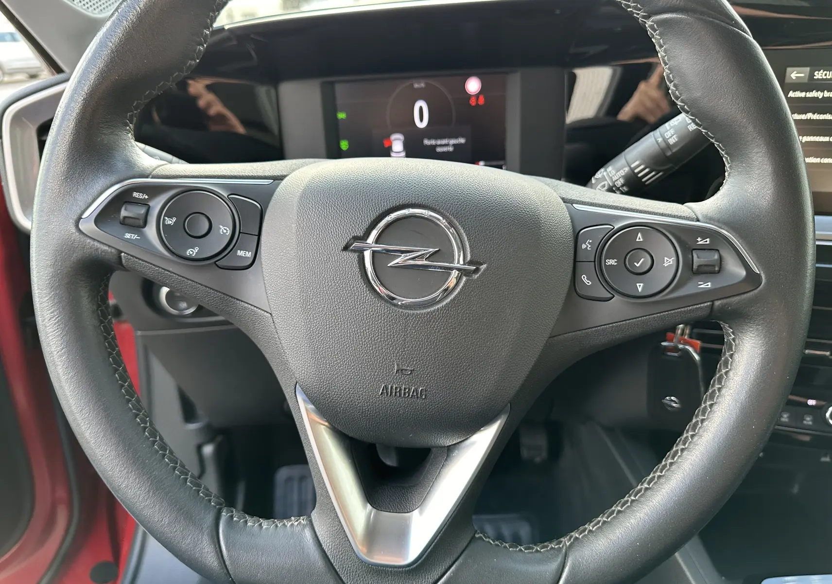 Gros plan sur le volant en cuir noir de l'Opel Mokka rouge Kardinal, avec commandes intégrées et tableau de bord numérique visible.