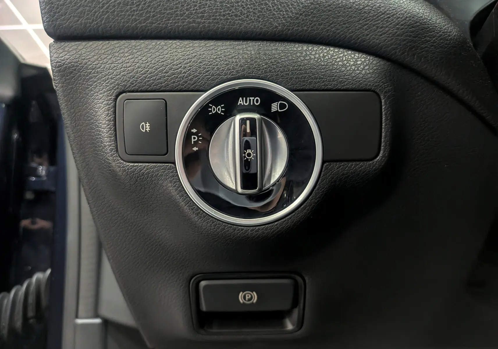 Gros plan sur le bouton de commande des phares et frein à main électrique dans une Mercedes Classe CLA 200 bleu Cavansite de 2017.