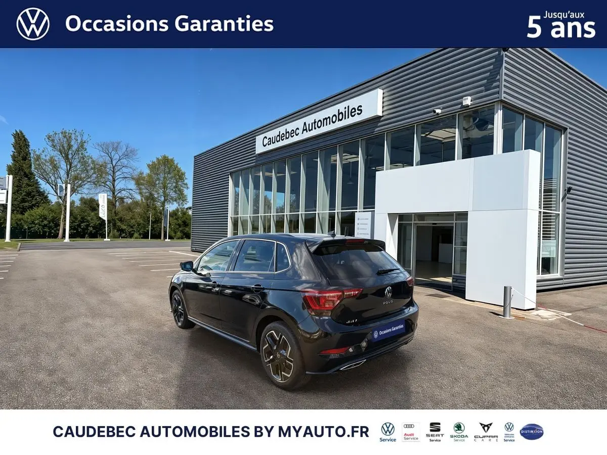 Vue 3/4 arrière droite d'une Volkswagen Polo noire intense nacrée, stationnée devant un bâtiment commercial moderne.