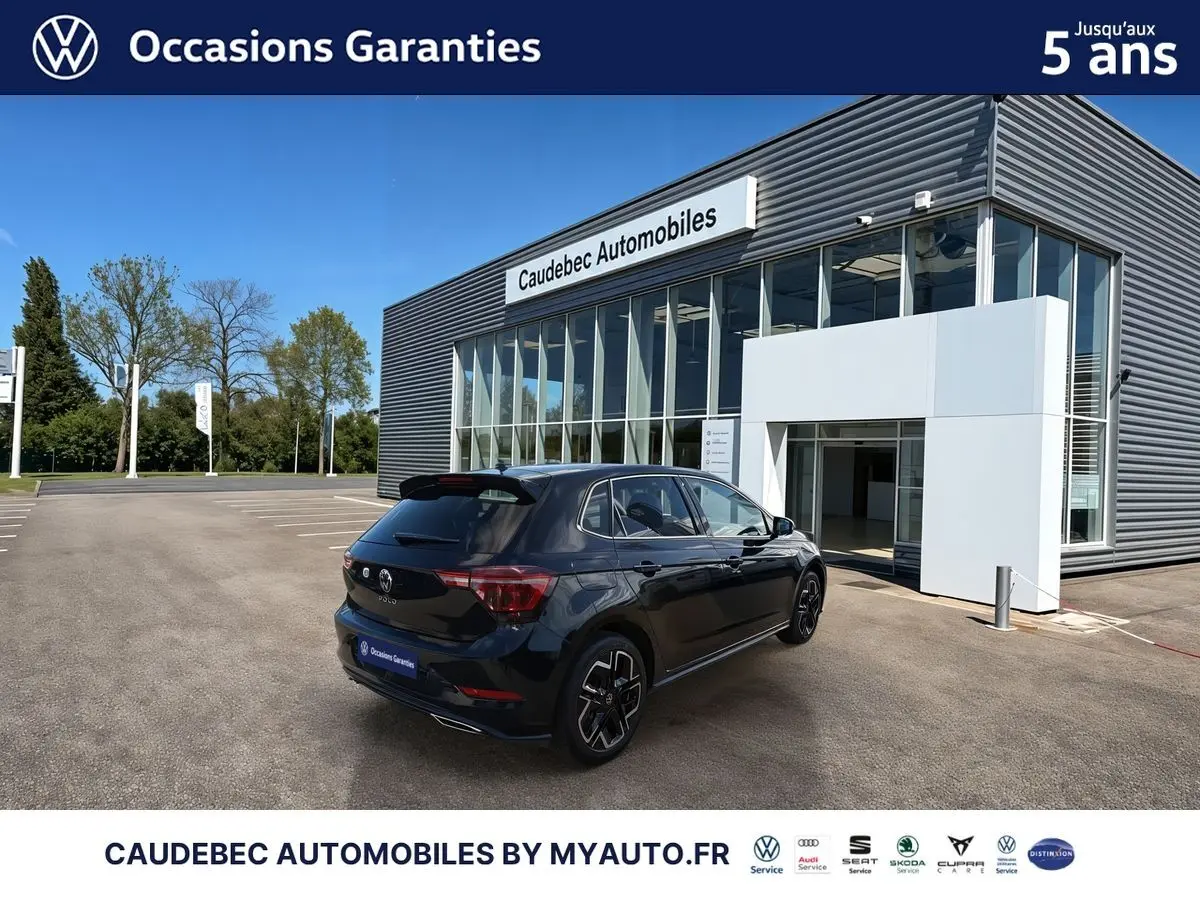 Vue 3/4 arrière droite d'une Volkswagen Polo noire intense nacrée stationnée devant un bâtiment moderne de concession.
