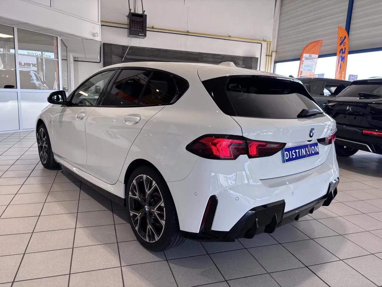 BMW Série 1 120d M Sport blanc vue 3/4 arrière droit en intérieur, avec jantes noires et détails sportifs.
