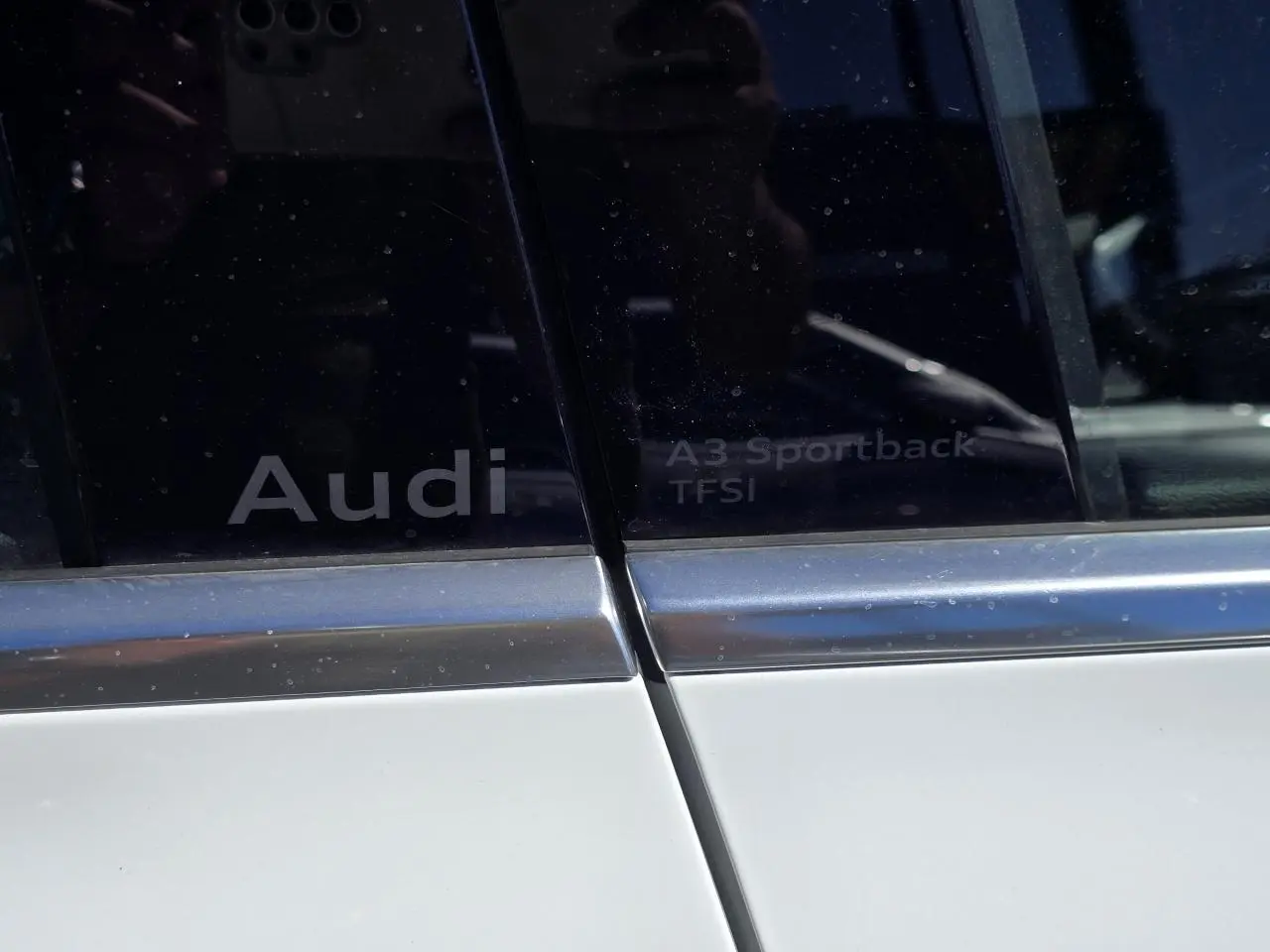 Gros plan sur les vitres arrière gauche d'une Audi A3 Sportback blanche avec logos Audi et TFSI visibles.