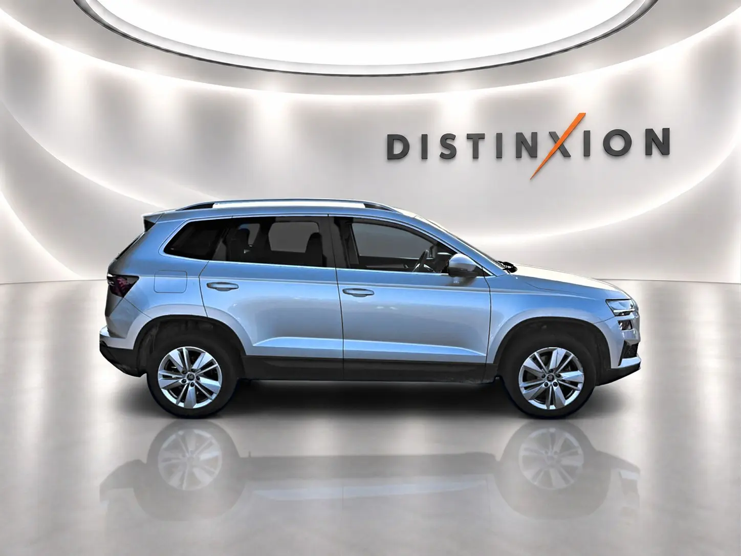 Vue de profil droit du SUV SKODA KAROQ gris clair avec barres de toit et jantes alliage, dans un showroom lumineux.