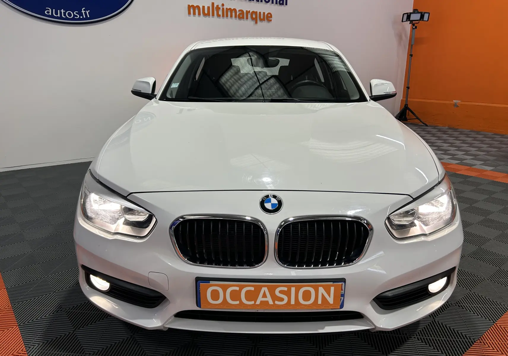 Vue avant d'une BMW Série 1 blanche 116d Business 5 portes avec phares et antibrouillards allumés en showroom.