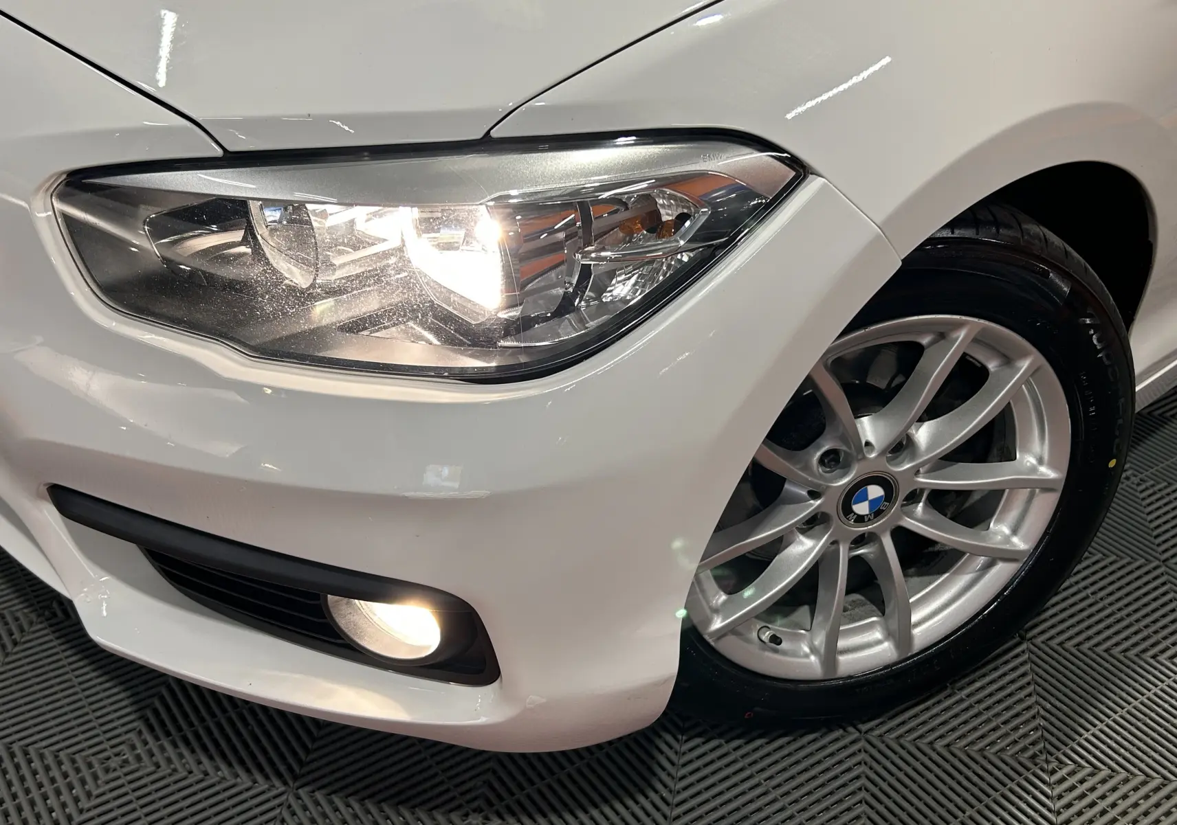 Gros plan sur l'avant droit blanc d'une BMW Série 1 116d avec phare allumé et jante alliage visible.