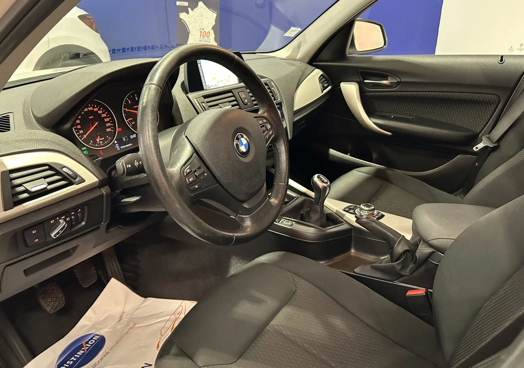Vue intérieure côté conducteur de la BMW Série 1 116d Business 2015 avec volant cuir, tableau de bord et levier de vitesse manuel.